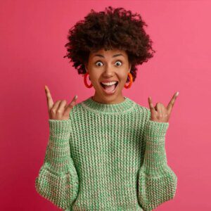 happy carefree dark skinned rebellious young woman enjoys awesome music makes rock n roll gesture has fun on music festival or cool event wears casual jumper poses against pink wall Website  design